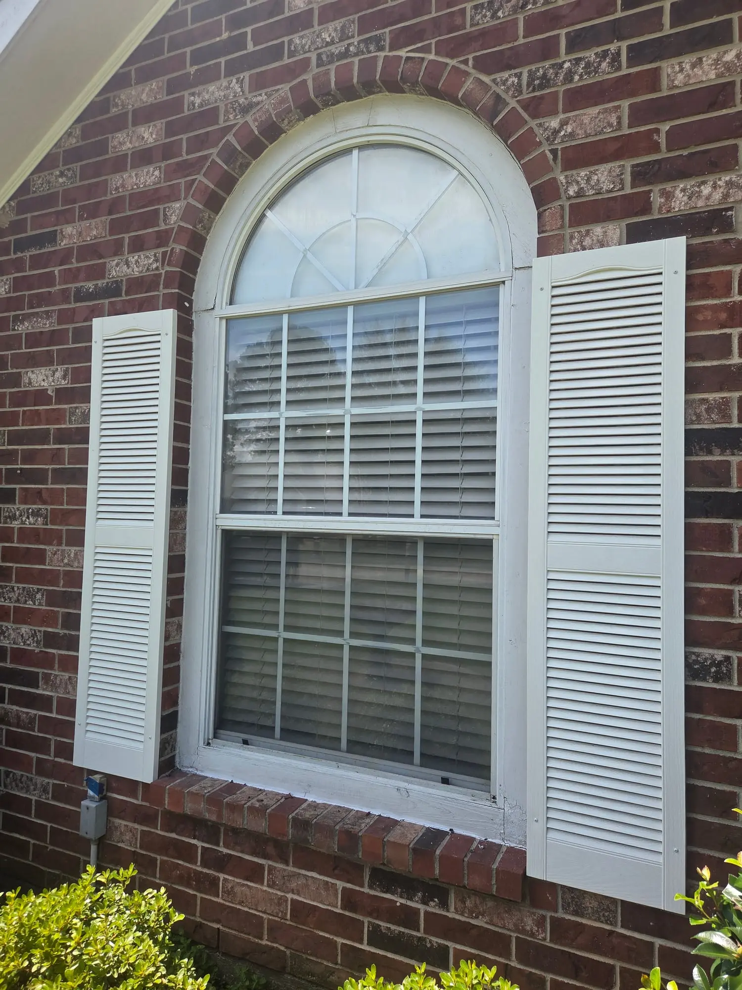 A clean arched double-hung window with white shutters on a brick home